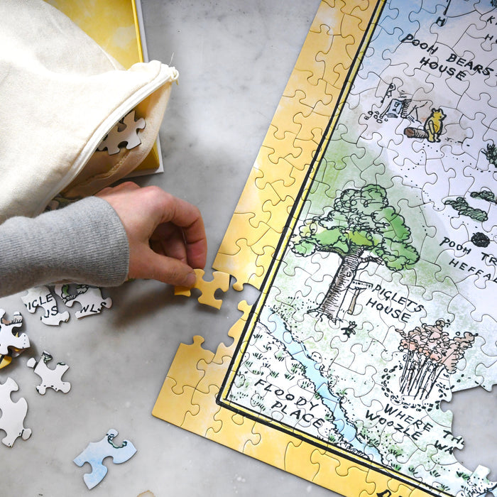 A person's hand placing a yellow border piece into a partially completed jigsaw puzzle of the Hundred Acre Wood map on a marble surface.