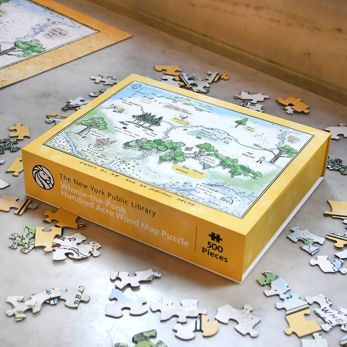 A yellow box for "The New York Public Library Winnie-the-Pooh Hundred Acre Wood Map Puzzle" sits on a marble surface surrounded by scattered puzzle pieces.