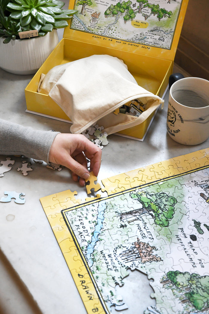 A person's hand picks up a yellow puzzle piece from a marble tabletop to place it into the edge of a partially completed Winnie-the-Pooh Hundred Acre Wood Map Puzzle.