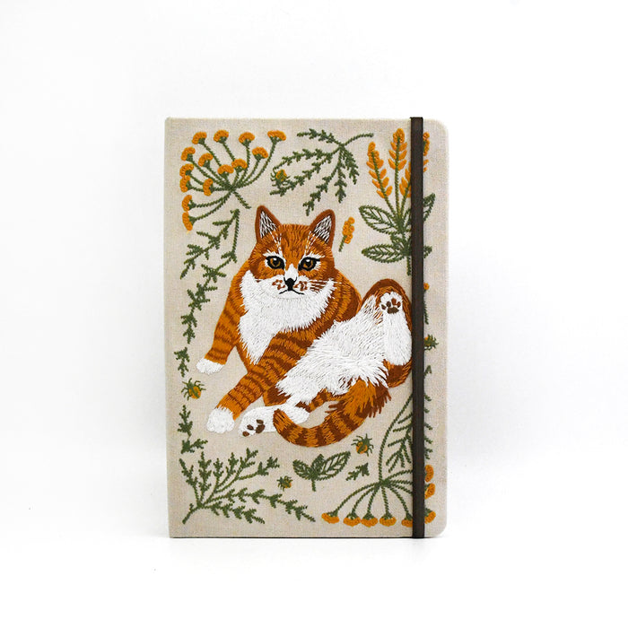 Notebook featuring an embroidered orange and white cat surrounded by green leaves and small yellow flowers on a beige background.