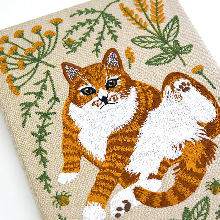 Close-up of notebook featuring an embroidered orange and white cat surrounded by green leaves and small yellow flowers on a beige background.