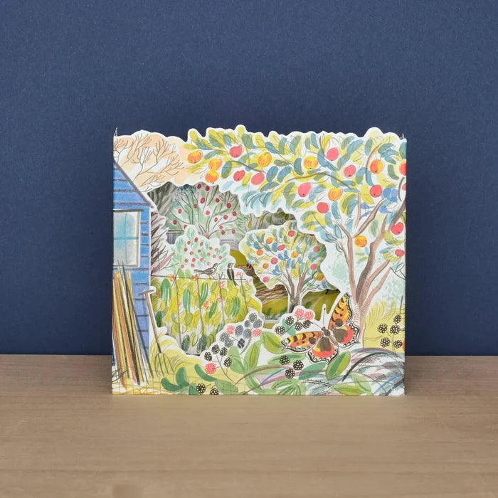 A straight-on front view of the 3D diorama die-cut card displaying intricate cut-out layers of an allotment garden with fruit-laden trees, plants, and a butterfly.