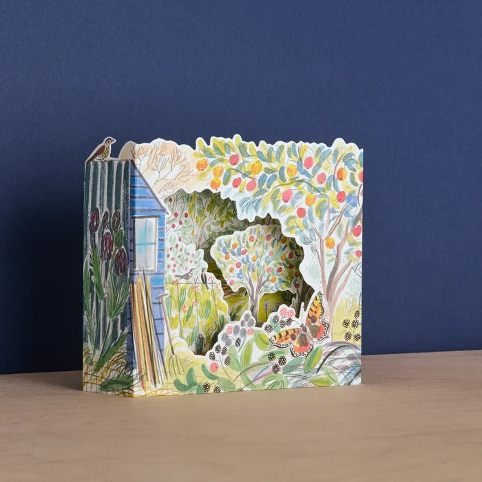 An angled view of a freestanding 3D die-cut card featuring layered illustrations of a garden orchard, a blue shed, fruit trees, and a butterfly.