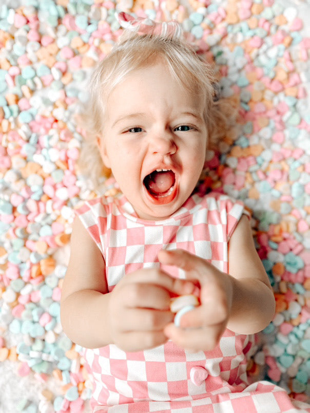 Toddler girl in pink checkered romper lying on pastel candy hearts – Adorable Valentine’s Day outfit for babies and toddlers. Soft and stylish checkered dress, perfect for festive photoshoots, playdates, and holiday celebrations. Cute and comfy handmade kids' fashion.