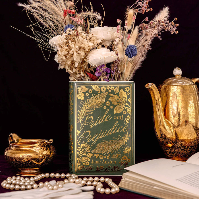 A decorative book-shaped vase featuring gold foil illustrations and the title 'Pride and Prejudice' on a dark green cover, filled with dried flowers.