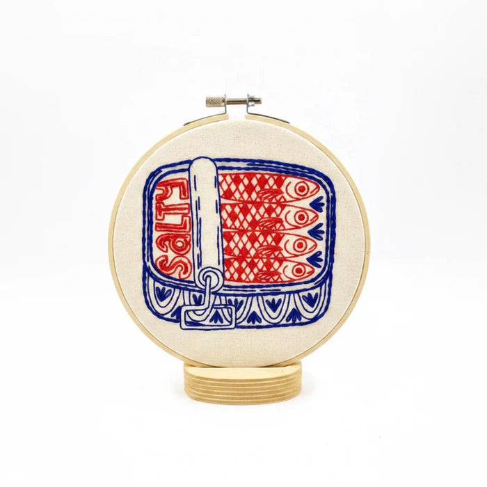 Embroidery hoop with half opened sardines tin in blue and red design on a white background