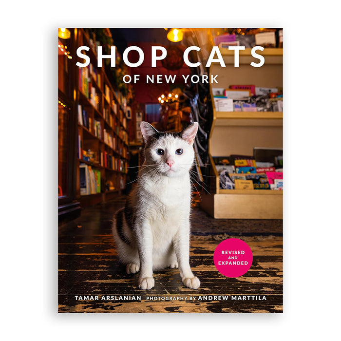 The book cover of 'Shop Cats of New York' by Tamar Arslanian, featuring a white and gray cat sitting on a wooden floor in a cozy, warmly lit bookstore. The title is written in bold white letters at the top, and a pink circular badge on the lower right reads 'Revised and Expanded'.