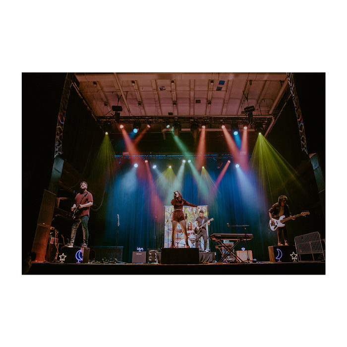 A band performs on stage under colorful lights, illuminated by glimmers spotlights. Four musicians play guitars and sing, surrounded by stage equipment and instruments against a dark background.