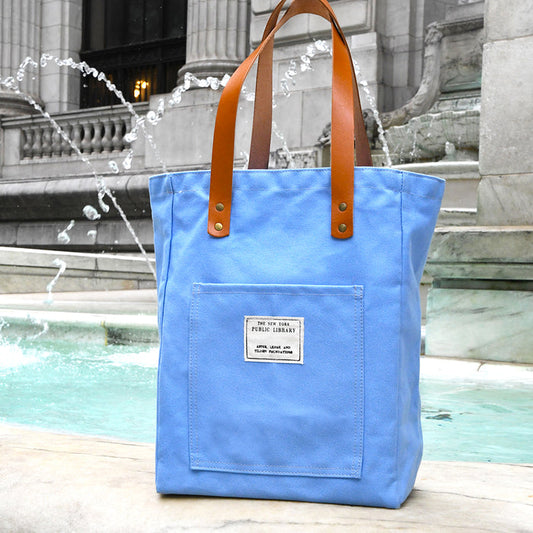 Sky blue tote bag with brown handles featuring a rectangular NYPL stamp on the front pocket placed in front of a fountain.