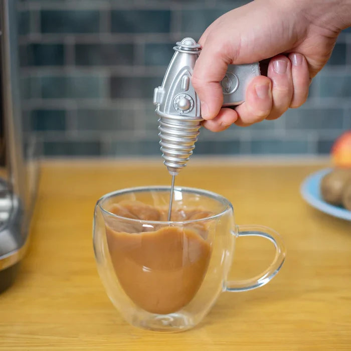 Person using a silver espresso frother shaped as a space weapon to stir coffee in a clear glass mug on a wooden table.