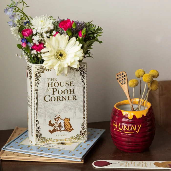 A styled tabletop scene with a white book-shaped vase featuring the cover of 'The House at Pooh Corner'. Vase is off white with gold detailings.