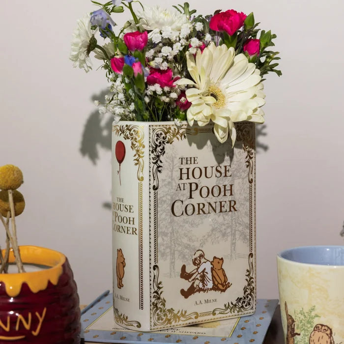 A decorative ceramic vase shaped like a vintage 'The House at Pooh Corner' book, filled with a bouquet of white, pink, and purple flowers, set on a table next to other ceramic mugs.
