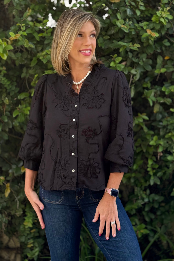 Woman wearing a black 3D floral textured blouse with 3/4 sleeves, ruffled neckline, and button-down front, styled with dark denim jeans, standing in front of greenery.