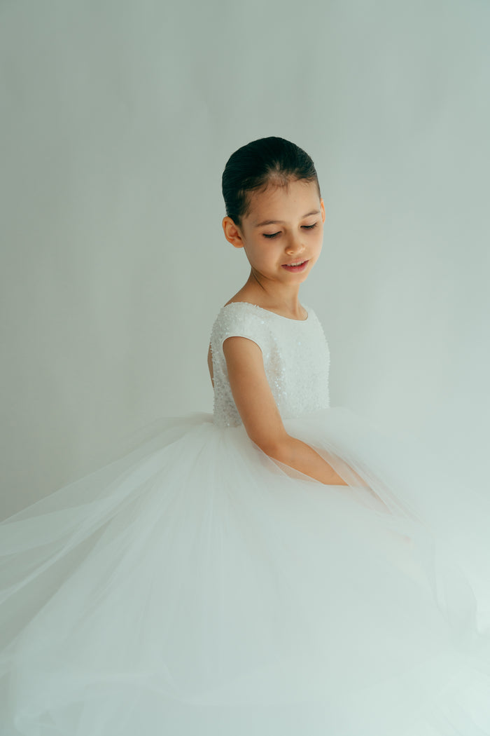 A young girl in a white, maxi dress adorned with sequins and beads, posing in a semi-profile stance.