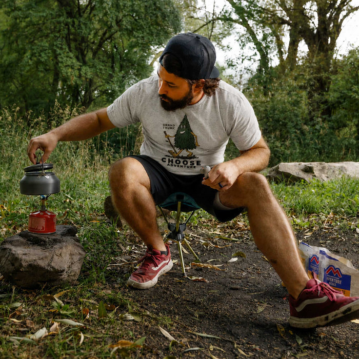 Man camping outdoors, using a portable stove with a scenic background.