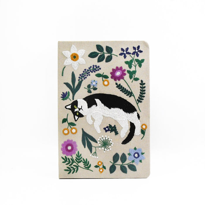 A journal with a beige cover, featuring an embroidered design of a black and white cat surrounded by colorful wildflowers and green leaves, shown against a plain white background.