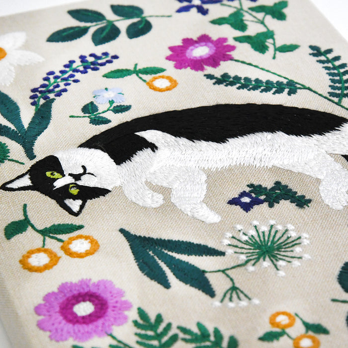 Close-up of a beige embroidered textile featuring a black-and-white cat surrounded by colorful floral motifs in green, purple, pink, blue, and orange.