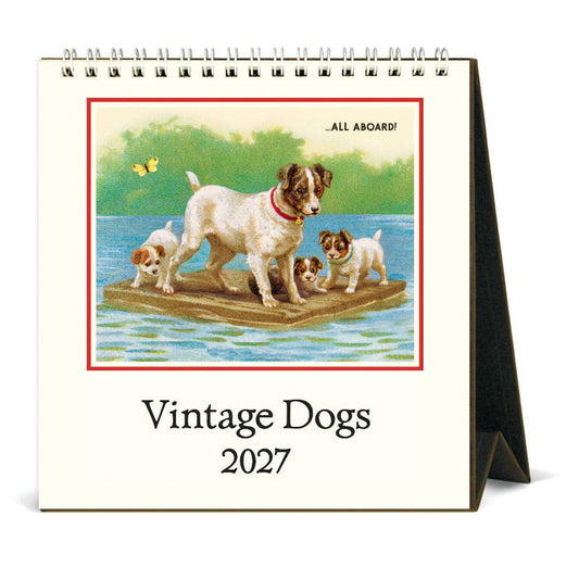 A spiral-bound desk calendar titled 'Vintage Dogs 2027' standing on a black easel stand. The front cover features a vintage illustration of several dogs on a wooden raft on water, with the text 'ALL ABOARD!' and 'Vintage Dogs 2027' below.