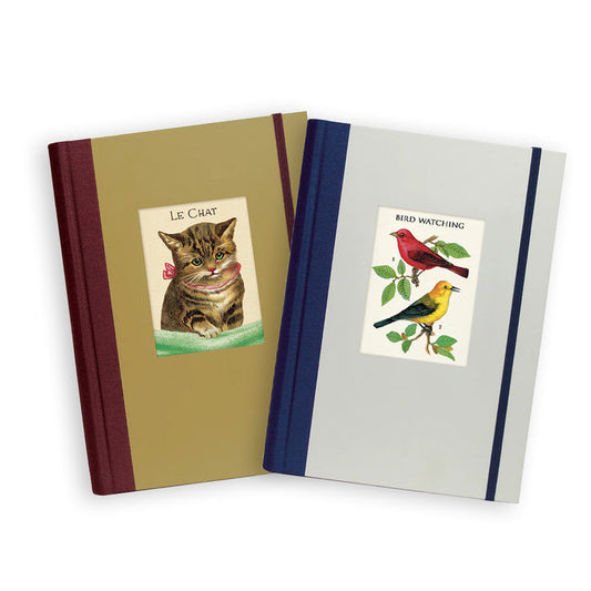Two hardcover notebooks or journals overlapping on a white background. The left journal has a tan cover with a dark red spine and a central label showing a tabby cat with the text 'LE CHAT'. The right journal has a light gray cover with a navy blue spice and a central label showing two wild birds with text "BIRD WATCHING".