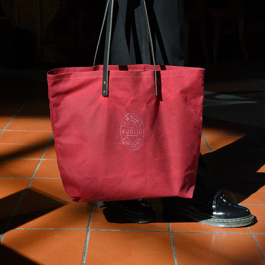 Red tote bag with a black leather straps featuring a NYPL oval logo.